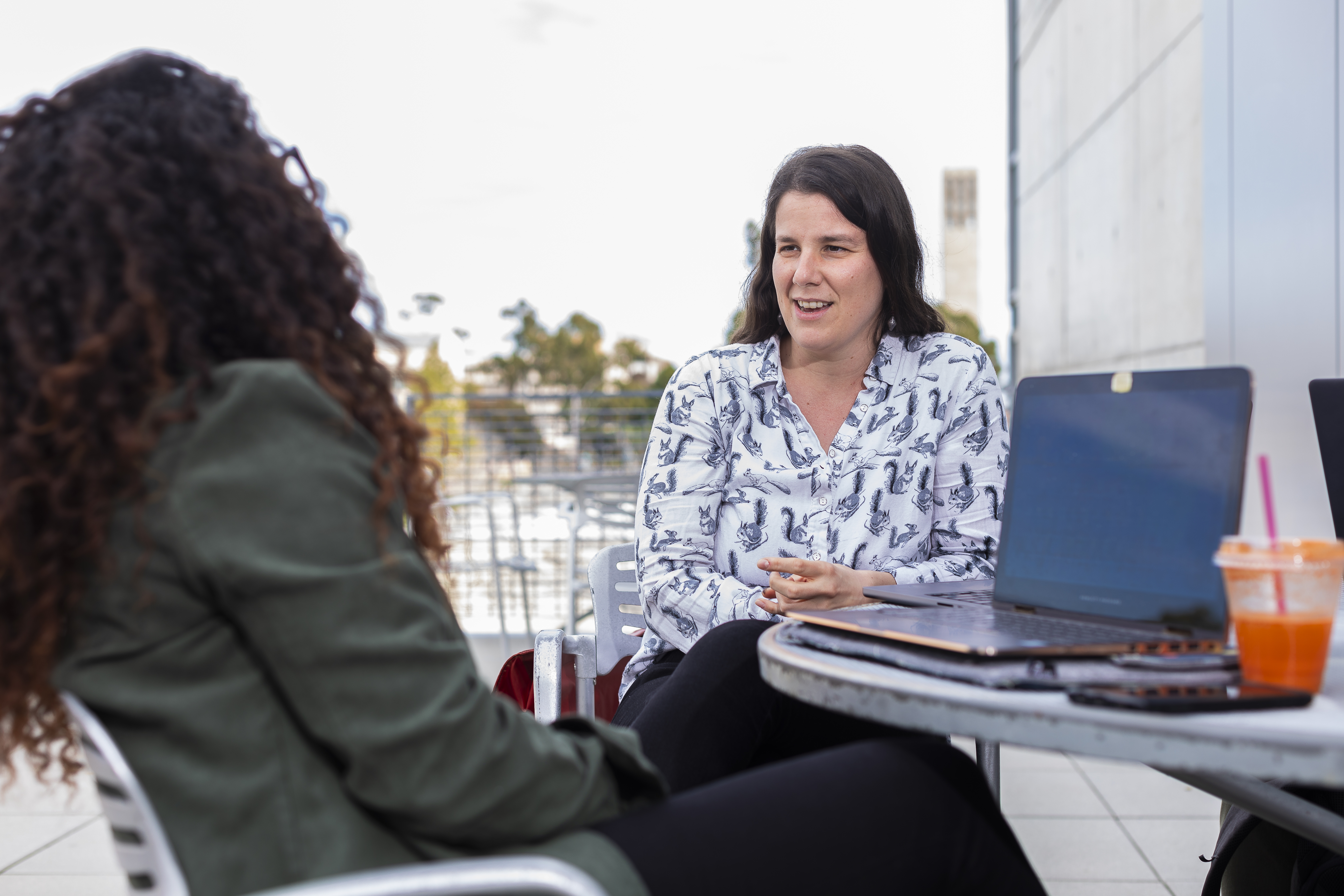 Two women sitting outside. One woman is facing away from the camera, the other is smiling and talking to her. 